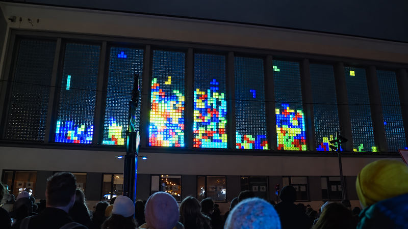 A crowd in winter hats watching falling-blocks games glow in a school’s glass-block windows at night