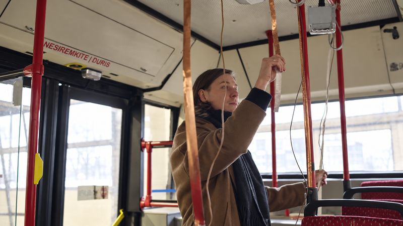 Inside the bus, a woman reaches up to a handrail wrapped in copper tape, cables hanging beside it