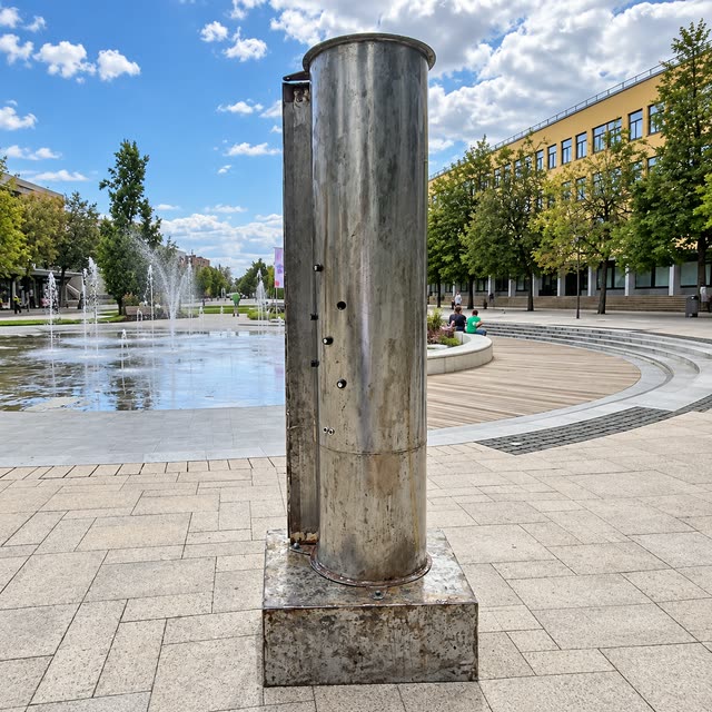 The Pipe Synthesizer, a metal pipe with knobs on a square base, standing on a paved city square with fountains behind it