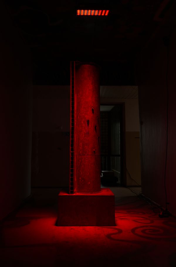 The Pipe Synthesizer, a tall metal pipe on a square base, standing in red light in a dark room